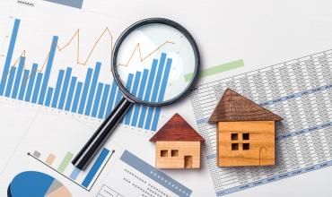 Two little wooden houses and a magnifying glass over papers with graphs and charts on them